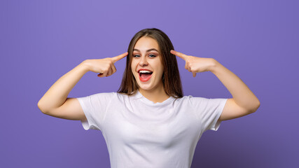 Eureka, finally got answer on question. Happy smiling girl pointing index fingers at her head, recalling or remembering, standing in blank white t shirt over purple background