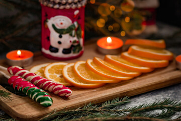 An orange cut into round slices lies on a wooden stand, next to orange candles and spruce branches