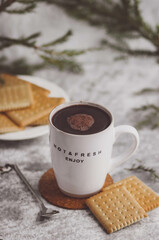 Light ceramic mug with coffee framed by green spruce branches, next to a plate with cookies