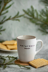 Light ceramic mug framed by green spruce branches, next to a plate with cookies. The inscription on the mug 