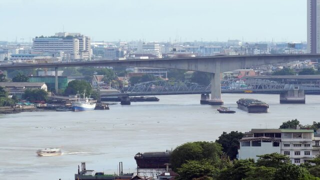 Train from three barges and tug-boats pass Chao Phraya River, move under Rama III and Krung Thep bridges, telephoto time lapse shot. Fast car traffic rush on roads, city buildings seen on background