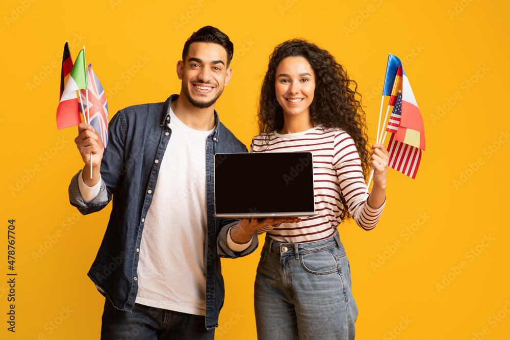 Poster smiling arab couple holding international flags and laptop computer with blank screen