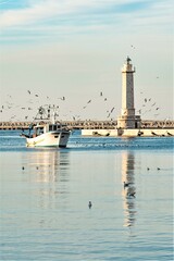 Fototapeta na wymiar Old fishing boat returning from fishing enters the port at sunset accompanied by a flock of seagulls. With beautiful lighthouse in the background, vertical