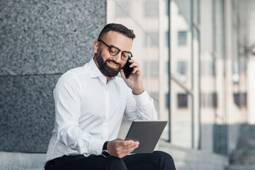 Handsome mature man having phone conversation, holding digital tablet, sitting near office center