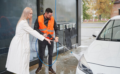 Young man washing car on carwash station outdoor, wearing orange vest. Handsome worker cleaning automobile, using high pressure water. Beautiful blonde woman client showing on the white car.