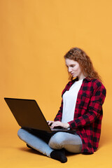 Naklejka premium red-haired girl in a plaid shirt sits with a laptop on an orange background