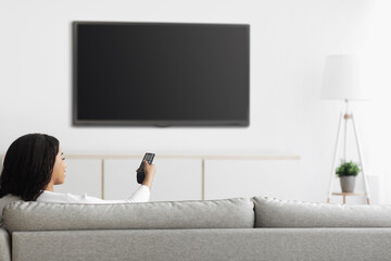 African american woman watching TV pointing remote control at flatscreen plasma television set with blank screen, mockup