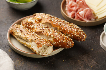 Savoury breakfast ingredients: brown lye bread sticks garnished with oats , pork slices prosciutto, hard cheese gouda and fresh greens on wooden boards