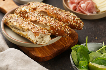 Savoury breakfast ingredients: brown lye bread sticks garnished with oats , pork slices prosciutto, hard cheese gouda and fresh greens on wooden boards