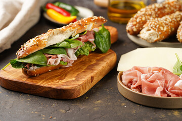 Long sandwich in brown lye bread sticks garnished with oats , pork slices prosciutto, hard cheese gouda and fresh greens on wooden boards