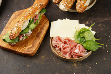 Long sandwich in brown lye bread sticks garnished with oats , pork slices prosciutto, hard cheese gouda and fresh greens on wooden boards