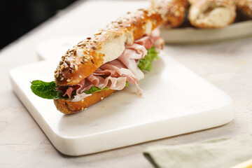 Long sandwich in brown lye bread sticks garnished with oats , pork slices prosciutto, hard cheese gouda and fresh greens on marble board, beige surface