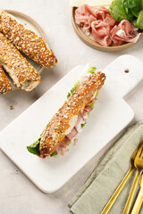 Long sandwich in brown lye bread sticks garnished with oats , pork slices prosciutto, hard cheese gouda and fresh greens on marble board, beige surface