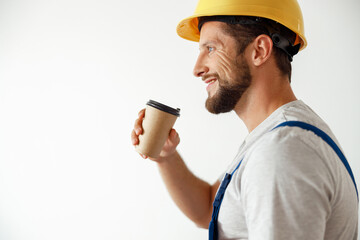 Side view portrait of smiling handyman in uniform and hard hat drinking coffee during a break while standing indoors