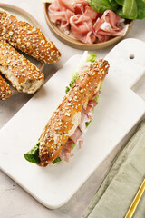 Long sandwich in brown lye bread sticks garnished with oats , pork slices prosciutto, hard cheese gouda and fresh greens on marble board, beige surface