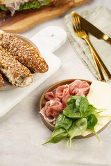 Savoury breakfast ingredients: brown lye bread sticks garnished with oats and seeds, pork slices prosciutto, hard cheese gouda on marble board