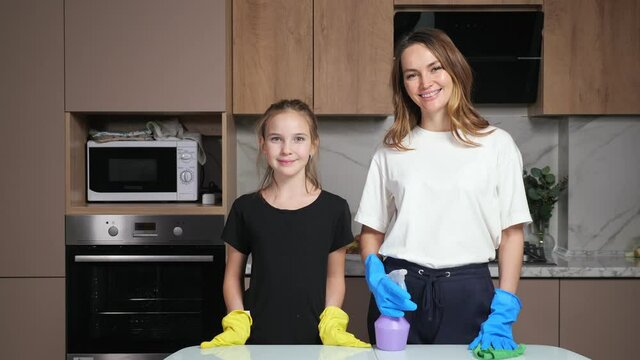 Mother and daughter with colorful gloves and cleanser stand near white table smiling and preparing for cleaning in kitchen of apartment.