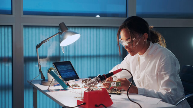 Close up of female electronics engineer in protective glasses checking motherboard with multimeter tester in advanced technology laboratory