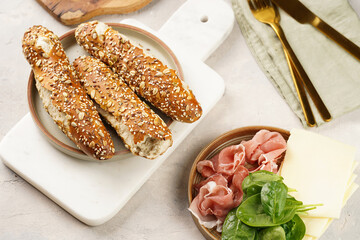 Savoury breakfast ingredients: brown lye bread sticks garnished with oats and seeds, pork slices prosciutto, hard cheese gouda and fresh spinach leaves on marble board