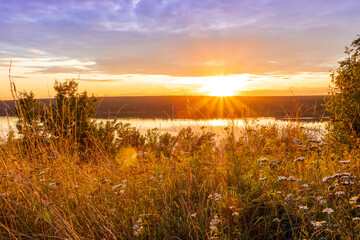 Amazing view at beautiful sunset or sunrise on a shiny lake from hill with green bushes, golden sun rays, calm water ,deep blue cloudy sky and forest on a background
