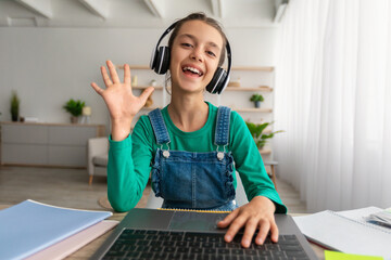 Girl sitting at table, using laptop, waving to webcam