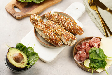 Savoury breakfast ingredients: brown lye bread sticks garnished with oats and seeds, pork slices prosciutto, hard cheese gouda and fresh spinach and avocado on marble board