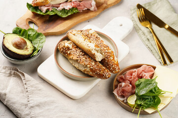 Savoury breakfast ingredients: brown lye bread sticks garnished with oats and seeds, pork slices prosciutto, hard cheese gouda and fresh spinach and avocado on marble board