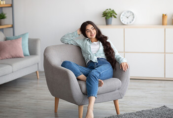 Fototapeta premium Gorgeous Indian woman sitting in comfy armchair, enjoying lazy morning, having peaceful weekend at home