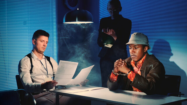 Medium Shot Of Black Prisoner In Handcuffs Having Interview With Caucasian Detective In Interrogation Room. Female Assistant Making Notes Of The Interrogation.