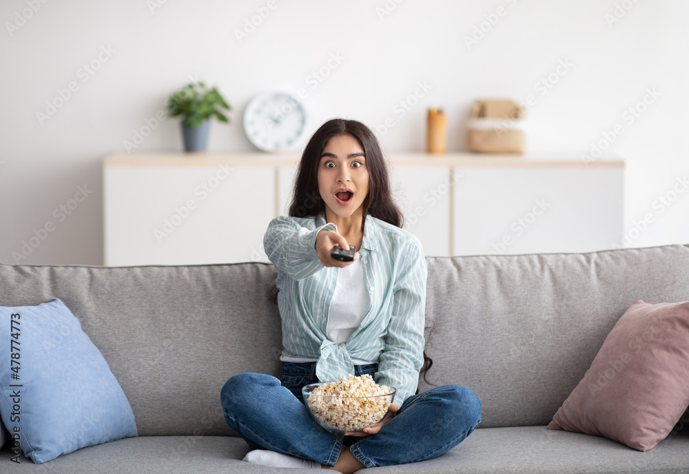 Wall mural full length of shocked indian woman sitting on sofa, eating popcorn, switching channels, watching tv