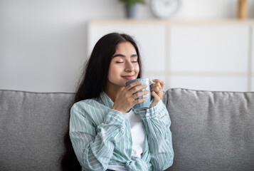 Portrait of beautiful Indian woman resting on couch, smelling aromatic coffee with closed eyes at home
