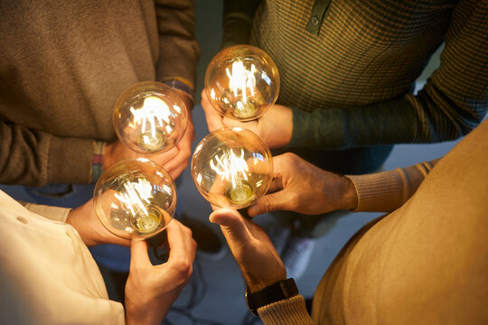 Team of four intelligent people together holding bright, shining, glowing Edison light bulbs as symbol of implementing innovations and developing modern, creative, clever ideas, close up, closeup shot