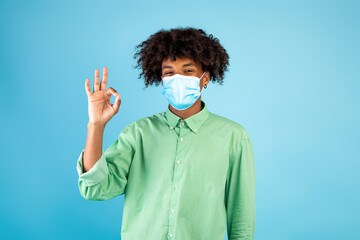 African american teen guy wearing protective face mask and gesturing okay sign, posing over blue background