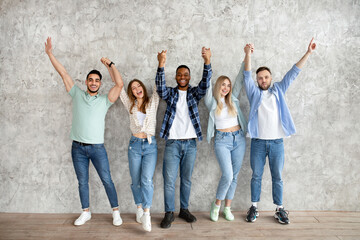 Obraz premium Full length of diverse friends holding hands up, feeling happy, looking at camera against grey studio wall