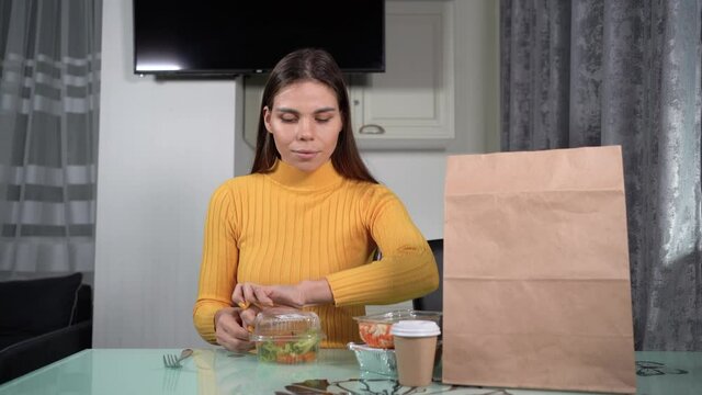 Caucasian woman sitting at home alone at the table opens a plastic disposable container with diet salad and starts having dinner. home delivery food concept.