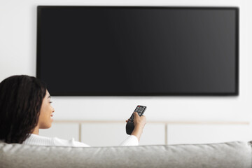 Modern technologies. Black woman watching TV pointing remote control at flatscreen television set...