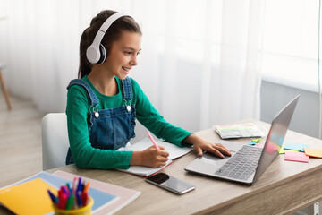 Girl sitting at desk, using laptop, writing in textbook