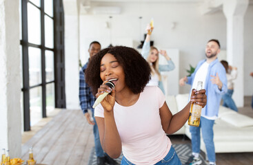 Happy black woman with bottle of beer and microphone performing song, singing karaoke on party with...