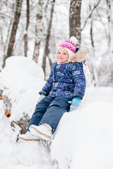 Little girl sitting on covered with snow tree log smiling looking at camera having fun wearing warm winter clothes in park. Awesome background full of white color.