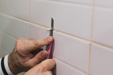 Close-up of man hands cutting extra part of dowels hammered into tiled wall in washroom with stationery knife. Renovation works in bathroom. Dirt from renovation process.