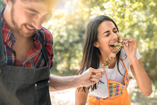 a young woman laughs as she eats skewers of meat from her friend's hands during a trip to the countryside. Lifestyle concept of young adults having fun spending time together with fun activities