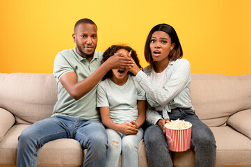 Scary moment in horror movie. African american parents closing daughter's eyes while watching TV, sitting on sofa