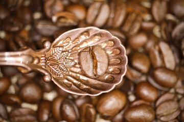 Top down view on isolated on vintage bronze spoon with one single isolated coffee bean, blurred beans background - obrazy, fototapety, plakaty