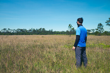 Naklejka na ściany i meble Young man standing in a deserted meadow He was looking out into the vast grassland. with a background of sky and pine trees., Rear view , man portrait on landscape , National Park ,Thailand