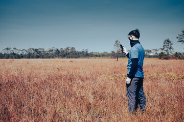 Naklejka na ściany i meble A young man wearing sunglasses and a windproof mask stands in a barren meadow. He was checking the weather forecast on his cell phone , side view , man portrait on landscape , National Park ,Thailand