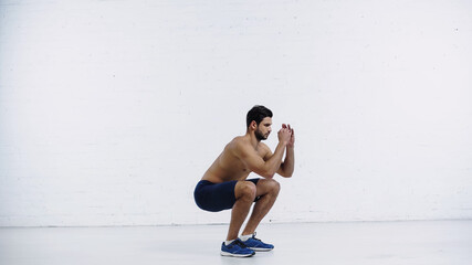 Obraz premium full length of young sportsman in shorts squatting near white brick wall