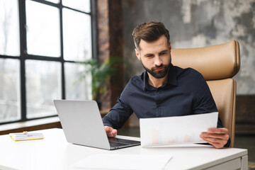 Worried caucasian man looking through correspondence. Guy holding documents in hands, got fine by post. Concentrated man sitting at the desk with a laptop in contemporary office