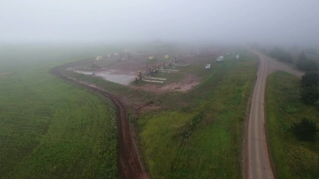 Aerial flying on oil pumps at oilfield cluster in a foggy field after rain. Muddy ground puddles hard working conditions.