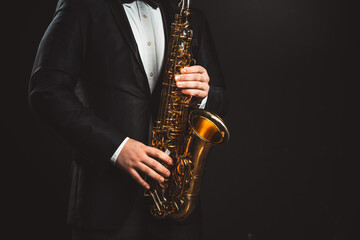 Fototapeta na wymiar Saxophonist Plays Jazz. Male Musician in a Formal Black Suit Holds a Tenor Saxophone on a Dark Background. Saxophone Close-up. Copy space High quality photo