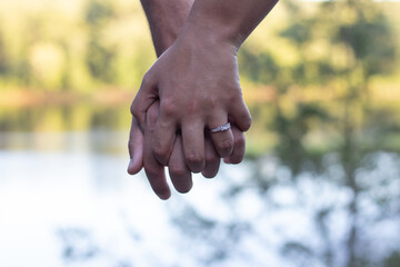 Couple's fingers entwined with a blurred background. Love is evident in the handhold. Sunny day background. Beautiful ring set on the hand. Forest leaves and water seen in the background. 